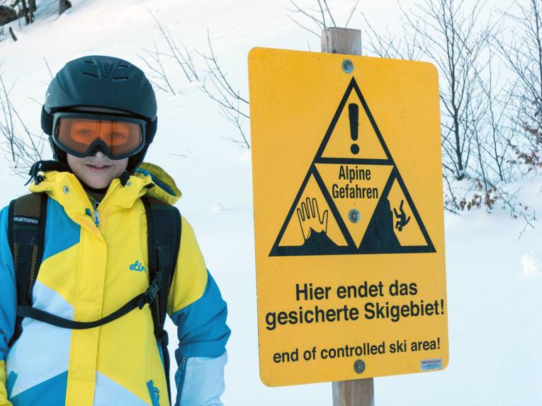 Ein Junge in Winterjacke und mit Helm auf dem Kopf steht neben einem gelben Schild. Auf diesem steht: Hier endet das gesicherte Skigebiet.
